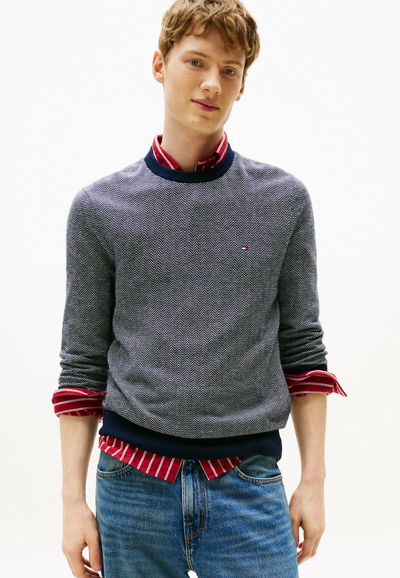 Maglione navy a trama, con motivo a spina di pesce, indossato sopra una camicia a righe rossa e bianca. Vestibilità slim e colletto, polsini e orlo a costine.