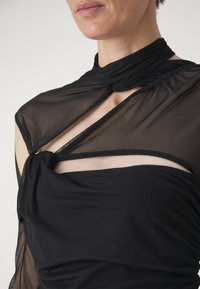 Rochie neagră cu panouri din plasă transparentă, accent legat la gât și material adunat. Textură fină cu decupaje strategice.