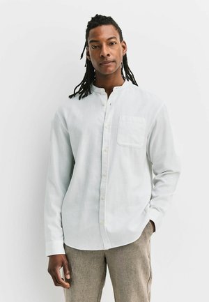 Mann mit langen Dreadlocks, trägt ein weißes Hemd mit durchgehender Knopfleiste und kragenlosem Ausschnitt sowie beige Hosen, steht vor einfachem weißem Hintergrund.