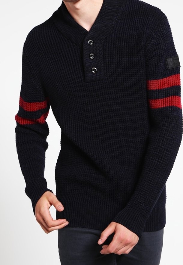Homme portant un pull en maille foncé avec trois boutons près du col et deux bandes rouges sur chaque manche sous l'épaule.