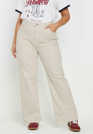 Pantalones de pierna ancha beige de algodón, con una cintura alta y trabillas para cinturón, combinados con una camiseta gráfica y zapatillas rojas.