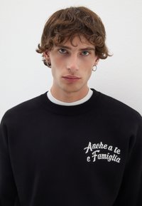 Felpa nera in morbido tessuto con testo ricamato bianco "Anche a te e Famiglia," e colletto a girocollo a coste.