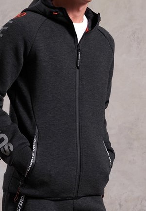 Mand iført en mørkegrå hættetrøje med lynlås og hænderne i lommerne, med sort lynlås og diskrete mærkede detaljer på ærmer og lommer.