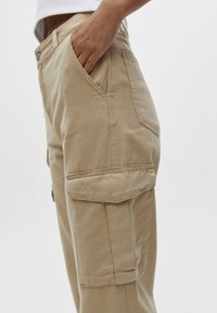 Persoon in beige cargobroek met zijflapzakken en hand gedeeltelijk in de voorkzak, tegen een effen lichte achtergrond.