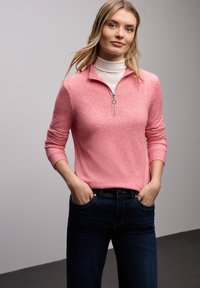 Różowy prążkowany sweter na zamek z długimi rękawami, zestawiony z ciemnoniebieskimi dżinsami. Sweter ma srebrny zamek błyskawiczny i wysoki kołnierz.