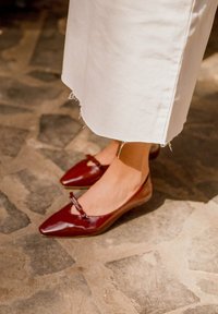 Burgundi patentinė oda balerinos bateliai su smailiu nosiu ir mažyte kaspino detale. Bateliai turi žemą kulną ir dėvimi su balta sijonu.