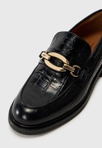 Svarte loafer i krokodillemønster med lær, dekorert med en gullfarget kjedeakcent, rund tå og svart teksturert såle.