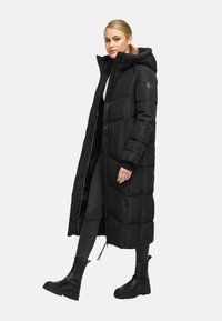 Femme portant un long manteau d'hiver noir rembourré avec capuche, un jean noir et des bottines noires, se tenant devant un fond clair et uni.