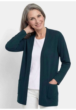Lächelnde Frau mittleren Alters mit grauen Haaren, die eine dunkelgrüne offene Strickjacke über einem weißen Hemd und weißen Hosen trägt, mit der Hand in der Tasche und den Hals berührend.