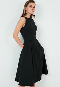 Egy ujjatlan fekete midi ruhát viselő nő zsebekkel, nagy fekete virágformájú fülbevalóval, egy sima háttér előtt áll.