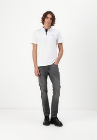 Camisa polo blanca de manga corta con un cuello azul marino, combinada con jeans de mezclilla gris y zapatos negros, de pie frente a un fondo blanco.