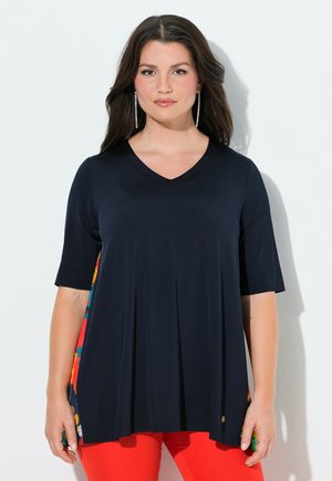 Kvinde med langt mørkt hår iført en marineblå V-hals top med blomstermønstrede sidepaneler og klare røde bukser, poserer foran en ensfarvet hvid væg.