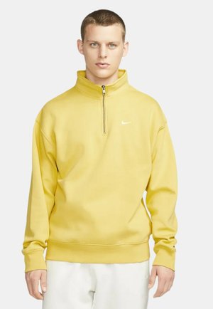 Geel Nike-pully met een hoge kraag en halve rits. Gemaakt van glad materiaal, met een klein wit logo op de borst.