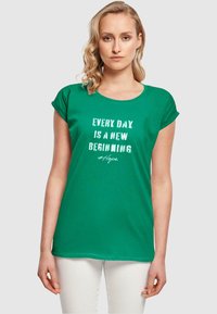Grünes Baumwoll-T-Shirt mit kurzen, aufgerollten Ärmeln, versehen mit weißem Text, der "Jeder Tag ist ein neuer Anfang #Hoffnung" liest. Glatte Textur.
