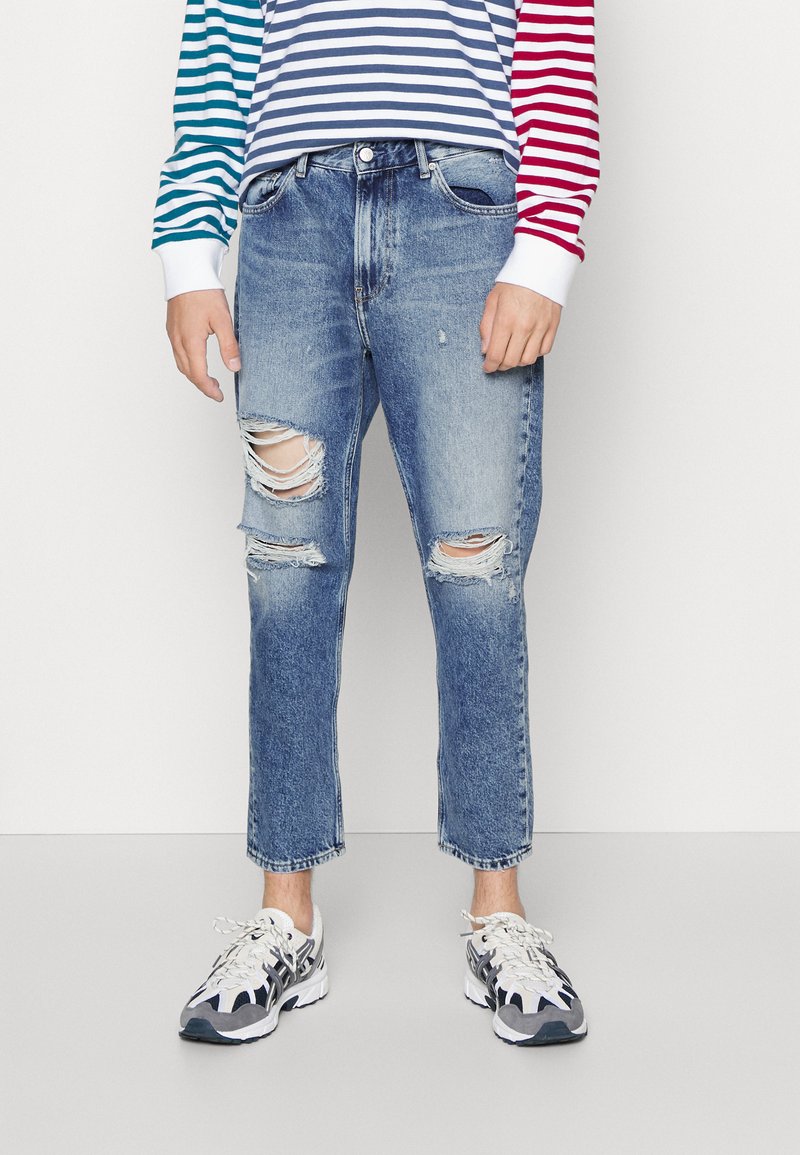 Slitte blå denimjeans med slitasjedetaljer, mid-rise liv, rett ben-kutt og lett falming. Kombinert med en flerfarget stripete skjorte og joggesko.