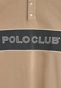 Beżowa koszulka polo z ciemnoszarym poziomym paskiem, na którym widnieje napis "POLO CLUB" w białym kolorze, z plakatką na dwa guziki i gładką teksturą.