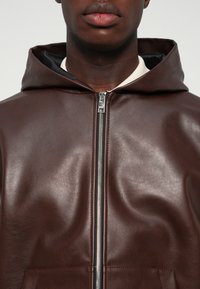 Gros plan sur une personne portant un sweat à capuche zippé en cuir marron foncé avec une doublure intérieure noire et une chemise blanche en dessous.