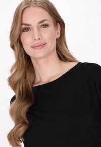 Schwarzes Oberteil mit weitem Ausschnitt, weicher Textur und enganliegenden Ärmeln. Die Haare sind wellig, und das Make-up ist natürlich mit neutralen Tönen. Der Hintergrund ist weiß.