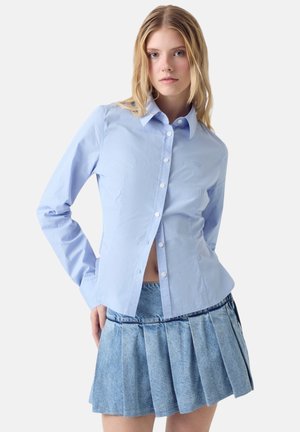 Giovane donna che indossa una camicia azzurro chiaro con bottoni e una minigonna di jeans a pieghe, in piedi con una mano dietro la schiena.