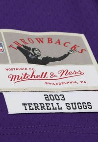 Lila Trikotstoff mit einem genähten Etikett, auf dem "Throwbacks," "Mitchell & Ness" und die Silhouette eines Spielers zu sehen sind. Zeigt "2003 Terrell Suggs".