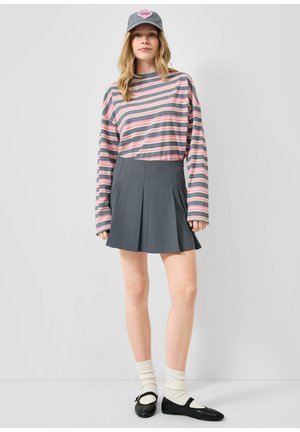 Jeune femme portant une chemise à manches longues rayée rose et grise, une jupe plissée grise, une casquette grise, des chaussettes blanches et des chaussures Mary Jane noires, debout.