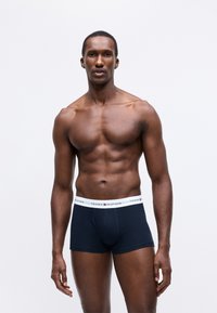 Muskuļots vīrietis stāv ar seju uz priekšu, valkājot tumši zilos Tommy Hilfiger boksierbikses ar baltu jostu uz vienkārša balta fona.