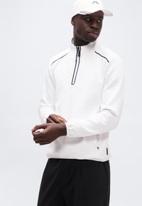 Άνδρας που φοράει λευκό πουλόβερ με φερμουάρ μέχρι τη μέση και λογότυπο Calvin Klein, λευκό καπέλο και μαύρο παντελόνι, προσαρμόζει το μανίκι, κοιτάζοντας προς τα αριστερά του.