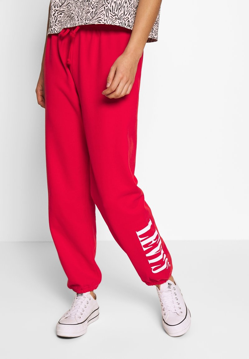 Personne portant un pantalon de survêtement rouge vif de Levi's et des baskets blanches, se tenant devant un fond blanc uni.