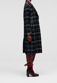 Cappotto a quadri in toni scuri con accenti rosa, abbinato a pantaloni lucidi bordeaux e stivali con tacco alto coordinati; trame ricche in tutto l'insieme.