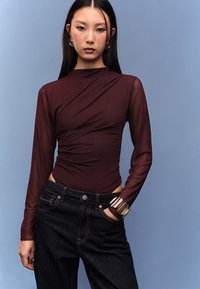 Viininpunainen pitkähihainen bodysuit, jossa on laskostettu muotoilu ja läpikuultavat hihat, yhdistetty tummiin denimfarkkuihin ja paksuun rannekoruun.