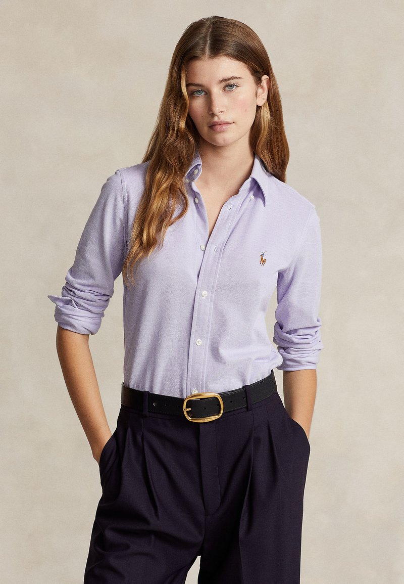 Light purple button-up shirt with rolled sleeves, featuring an embroidered logo. Paired with high-waisted dark pants and a black belt.