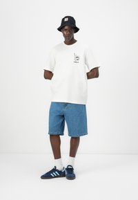 Hvit bomulls t-skjorte med grafikk, blå denimshorts, svart bøttehatt med logo, og blå joggesko med lyseblå striper.