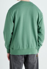 Sudadera verde con cuello redondo y mangas largas, con puños y dobladillo acanalados. Combinada con vaqueros gris oscuro.