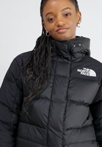 Crna izolirana puffer jakna s visokim ovratnikom, s logotipom The North Face na prsima i kvadratičastim uzorkom za dodatnu toplinu.