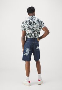 Shorts in denim blu scuro con ricamo di tigre sulla gamba sinistra; camicia a maniche corte con stampa tropicale in tonalità di nero e verde; sneakers bianche.
