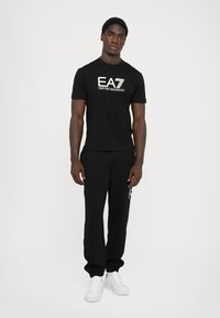 Camiseta negra con logo blanco de EA7 Emporio Armani; combinada con pantalones de chándal negros y zapatillas blancas. Diseño sencillo, tejido de algodón, corte casual.