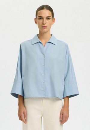 Vrouw draagt een lichtblauw oversized overhemd met brede mouwen en crèmekleurige broek, staand tegen een effen lichte achtergrond.