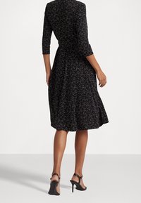 Svart klänning med ett mönster av små vita och gyllene prickar, trekvartsärmar och midjefokus, kombinerad med svarta högklackade sandaler.