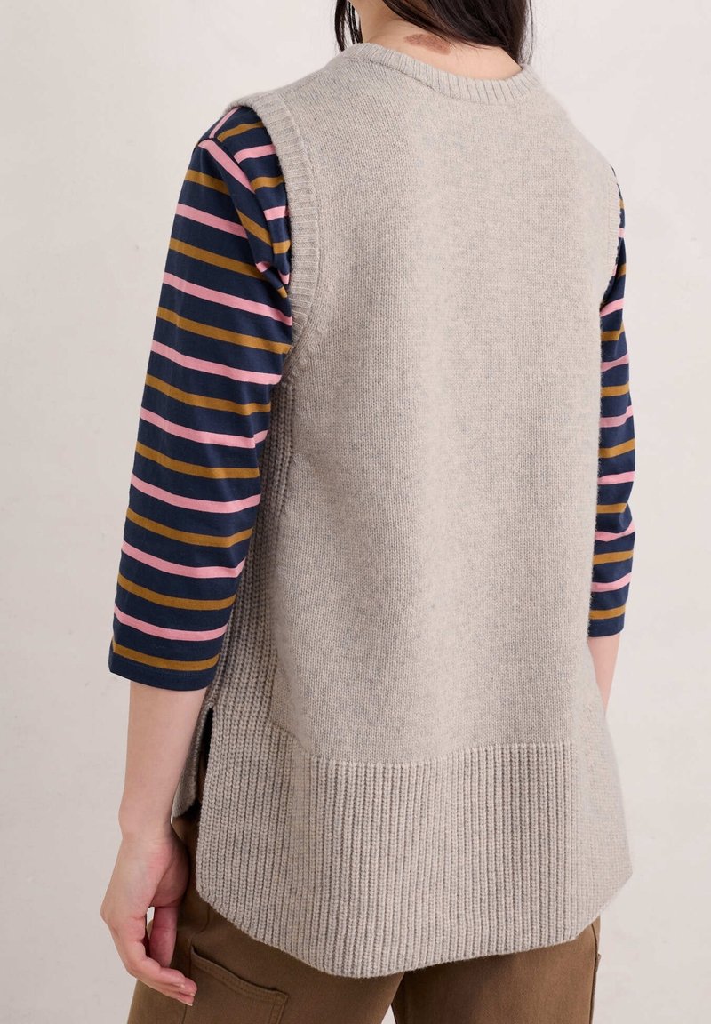 Beige ärmelloser Pullover mit geripptem Saum, getragen über einem gestreiften Langarmhemd in Marineblau mit pinken und goldenen Streifen.
