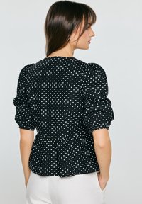 Mujer con blusa negra con lunares blancos y mangas abullonadas tres cuartos, de pie con las manos en los bolsillos de pantalones blancos, de perfil.