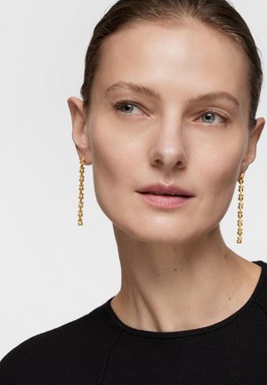 Primer plano de una mujer que lleva pendientes largos de cadena dorada, mirando hacia su izquierda, con el cabello recogido y vestida con una camiseta negra.