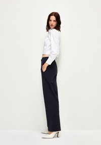Gekürzte weiße Bluse mit langen Ärmeln, kombiniert mit marineblauen Nadelstreifen-Hosen und spitzen weißen Schuhen mit schwarzen Akzenten.