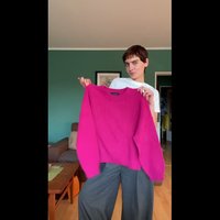 Leuchtend pinkfarbener gerippter Pullover mit langen, lockeren Ärmeln und Rundhalsausschnitt, gehalten vor einer grünen Wand. Darunter werden graue weit geschnittene Hosen getragen.