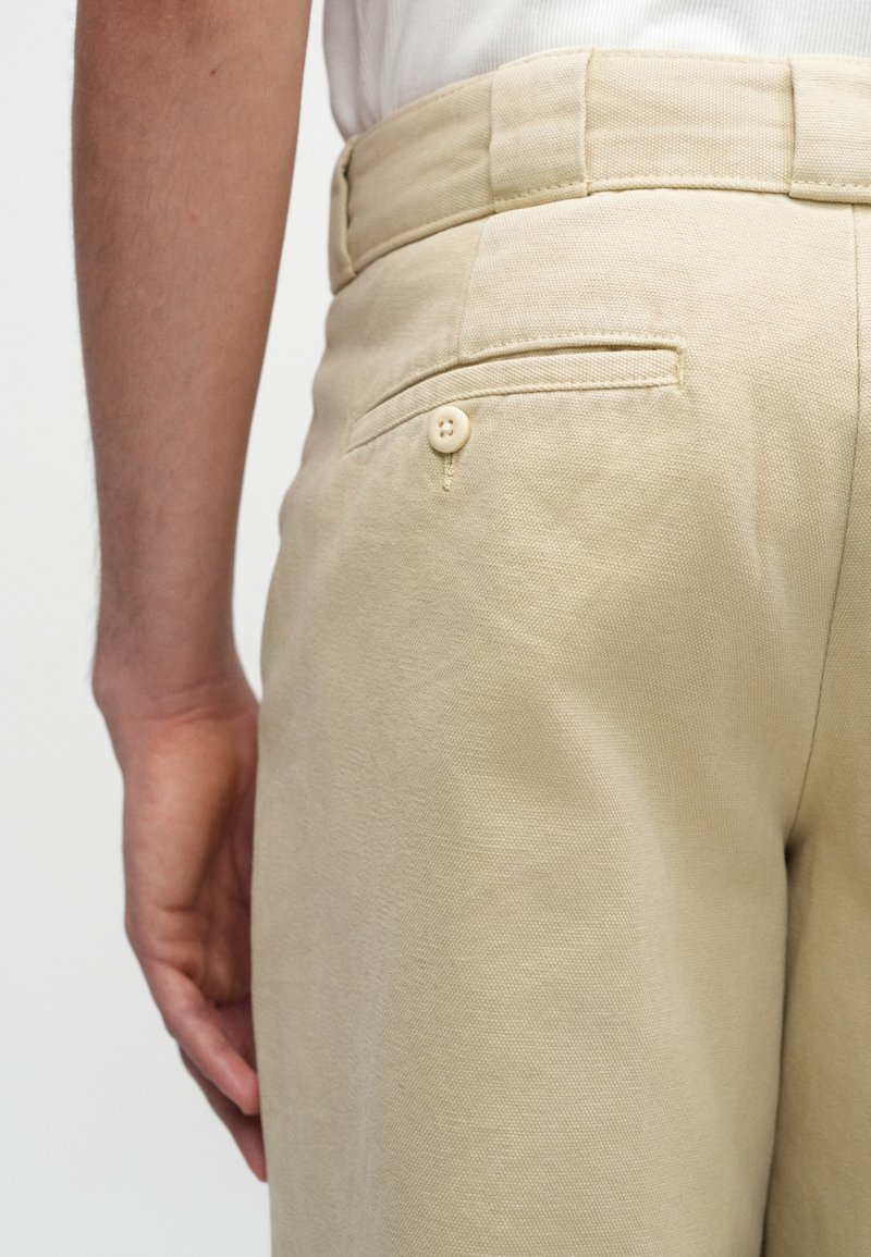 Pantaloni di cotone beige con una tasca posteriore, dotati di chiusura a bottone. La texture è liscia e presenta una finitura pulita e sartoriale.