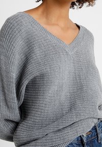 Femme portant un pull en maille gris clair à col en V ample et un jean bleu, mettant en valeur le tissu texturé et la coupe décontractée.
