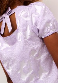 Donna che indossa un top in broccato lilla con maniche a palloncino e un dettaglio di legatura sul retro che crea un'apertura a forma di chiave.
