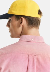 Rosa Hemd mit Button-Down-Kragen und Faltendetail am Rücken. Getragen mit einer gelben Mütze mit dunkem Schirm.