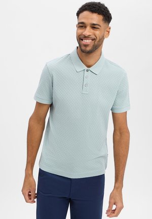 Lächelnder Mann, der ein hellblaues, strukturiertes Kurzarm-Poloshirt und dunkelblaue Hosen trägt, steht vor einem schlichten hellen Hintergrund.