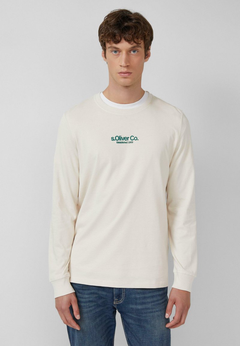 Long-sleeve cream T-shirt with ribbed cuffs, featuring green embroidered branding on the chest. Paired with blue jeans.