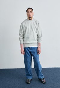 Sudadera de felpa gris con puños acanalados, llevada sobre una camisa blanca. Pantalones vaqueros azules holgados, usando zapatos negros y verdes, de pie sobre una alfombra.